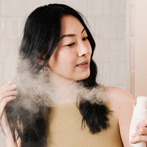 A woman with long black hair wearing a beige ribbed top is shown from the chest up, holding a white spray bottle while mist is sprayed onto her hair and neck. The background shows light beige tiled walls.